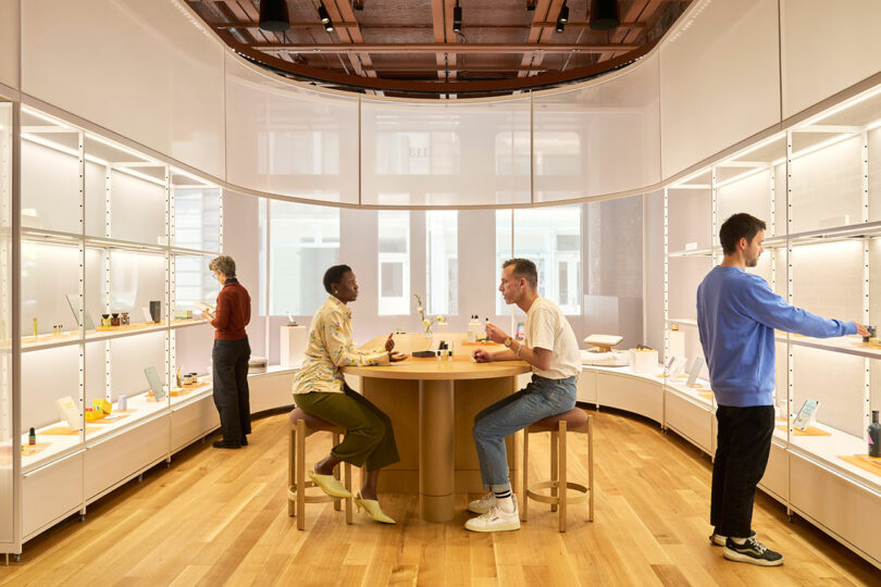 Four people interact with products in a modern, well-lit retail store with wooden floors and shelves displaying various items. Two people sit at a round table in the center.
