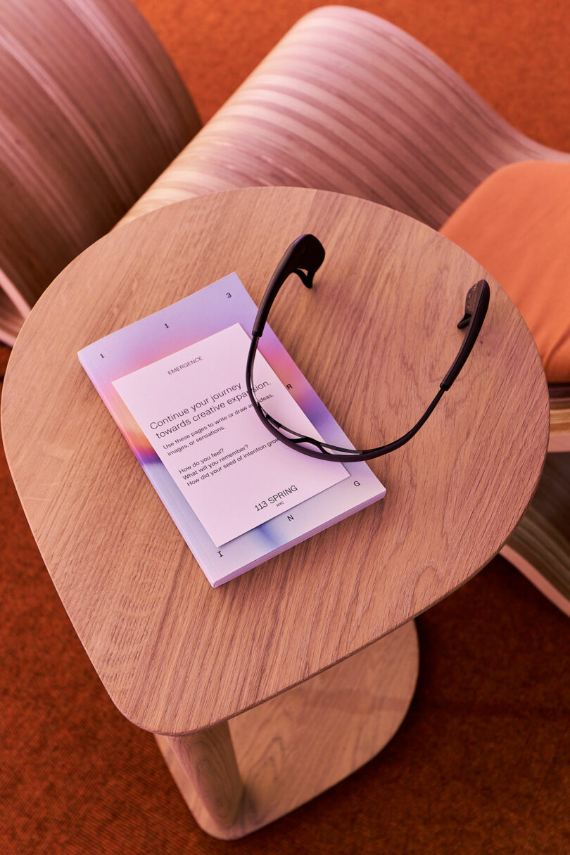 A pair of black glasses rests on a light wooden table next to a booklet with text, beside a curved wooden chair with an orange cushion, on an orange carpeted floor.