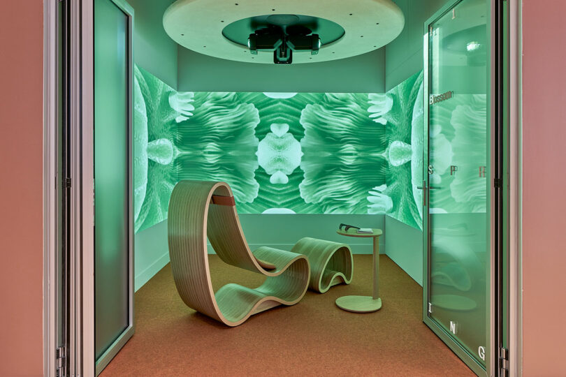 A modern relaxation room with a wavy wooden lounge chair, small side table, and walls displaying green abstract digital art.