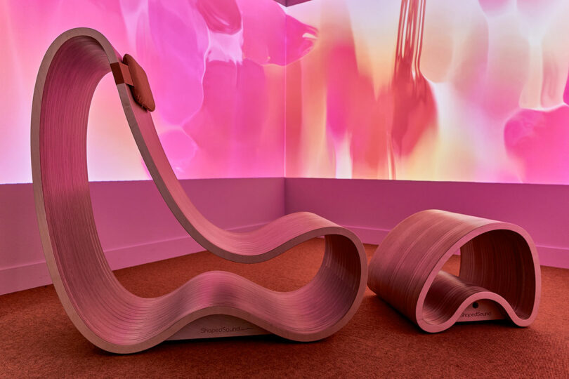 Curved wooden lounge chair and footrest in a room with pink and orange abstract projected light patterns on the walls.