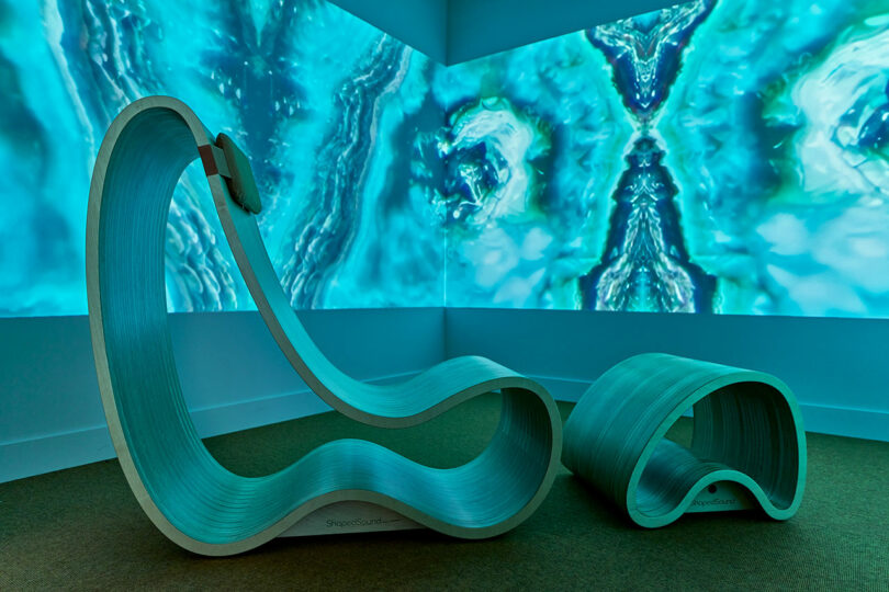 Modern, sculptural lounge chair and ottoman in a room with abstract blue and green digital art projected on the walls.