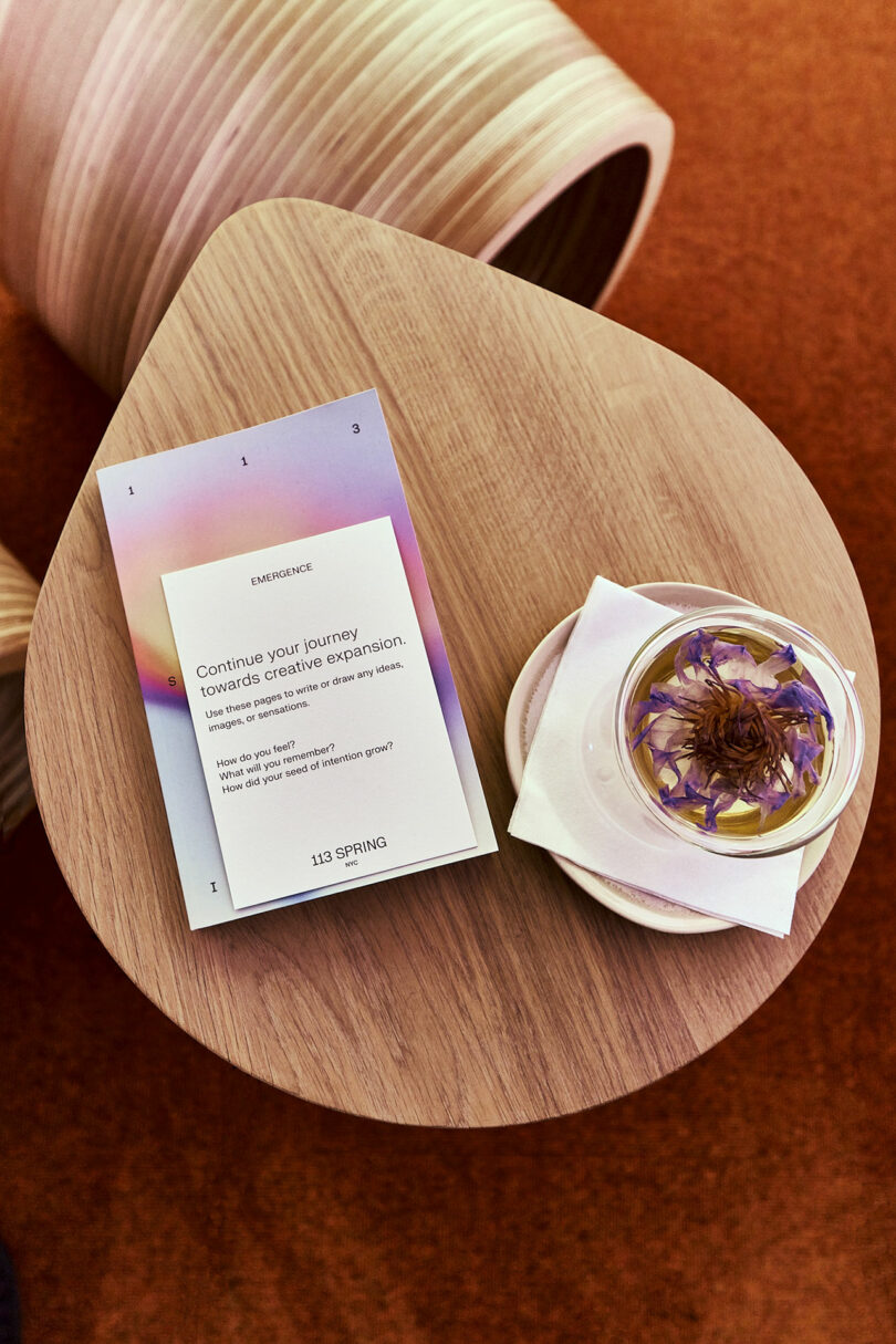 A small wooden table with a card displaying motivational text and a glass cup garnished with flower petals on a saucer, set on a reddish-brown carpet.