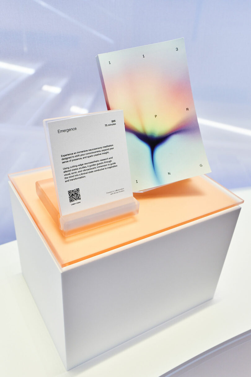A book titled "Emergence" with a pastel abstract cover is displayed upright on a lit pedestal next to an information card with a QR code.