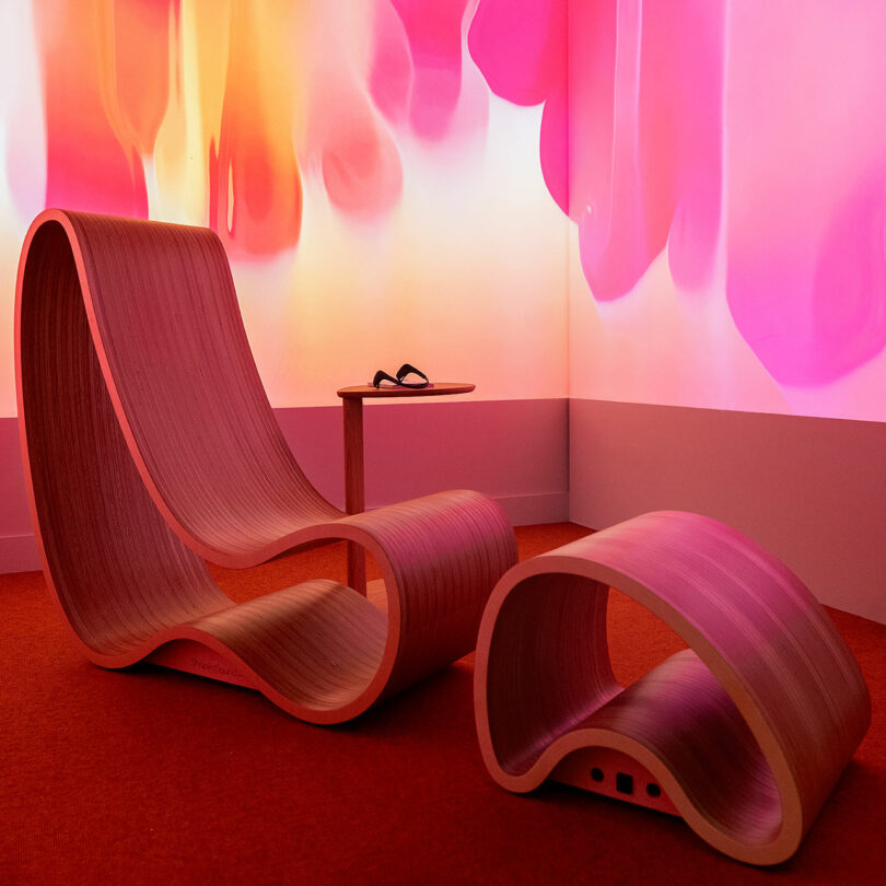 Curved wooden lounge chair and matching footrest on red carpet, with a small side table and pink abstract wall projection in the background.