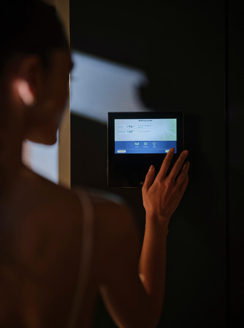 A person adjusts settings on a digital control panel mounted on a dark wall, with information displayed on the screen.