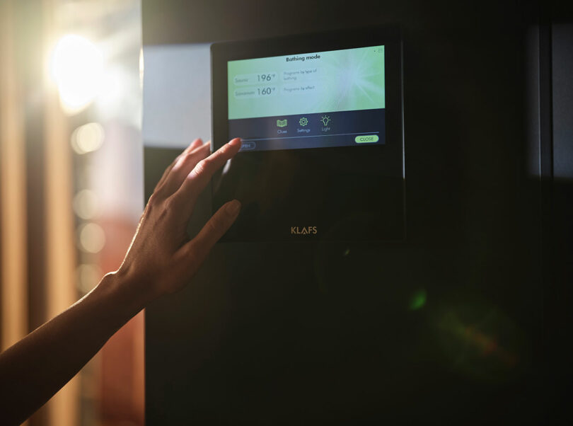 A person’s hand adjusts a touchscreen control panel displaying sauna settings on a device labeled “Klafs.”.
