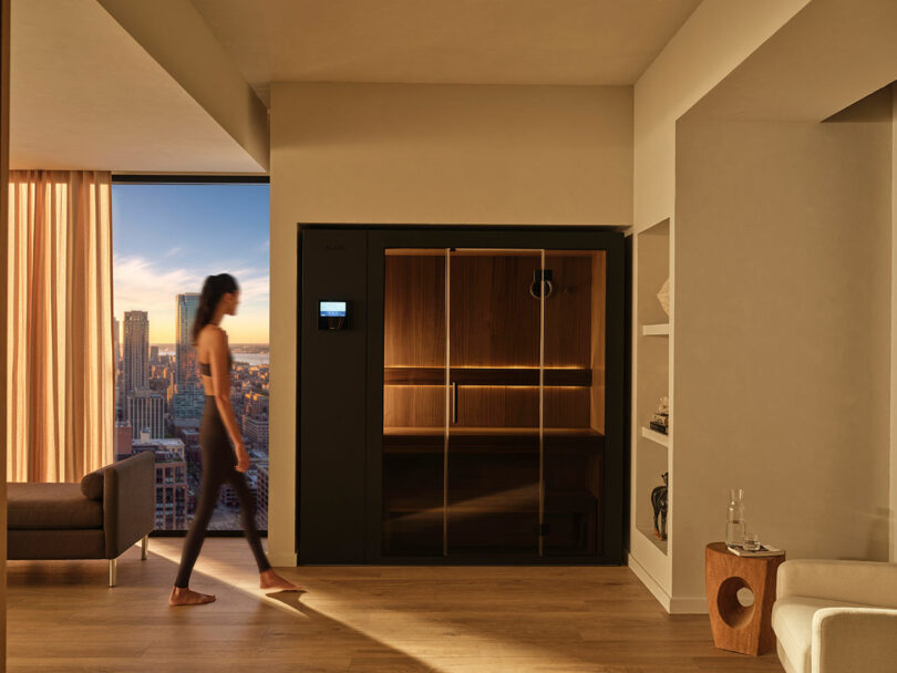 A person walks through a modern living room with wood flooring, large windows showing a cityscape, and a built-in sauna with glass doors.