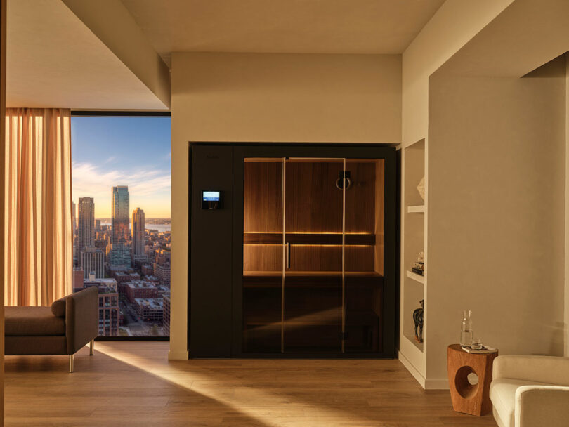 Modern apartment interior with a wooden sauna unit, floor-to-ceiling window showing a city skyline at sunset, and minimal furniture including a sofa and small side table.