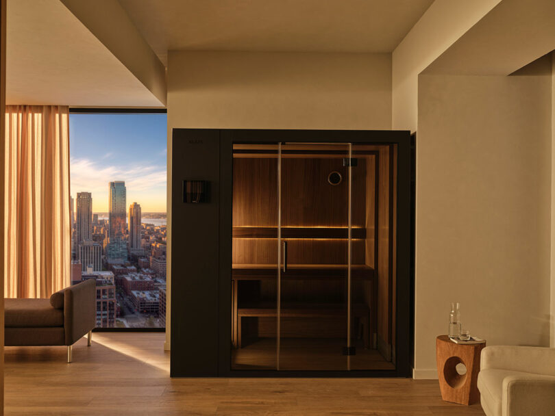 A modern indoor sauna with glass doors is situated in a minimalist room with wooden floors and large windows overlooking a cityscape at sunset.
