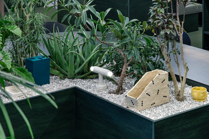 A variety of green indoor plants are arranged in a raised garden bed with white gravel and several geometric ceramic sculptures.