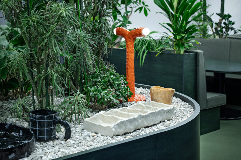 A small indoor garden with various green plants, white gravel, ceramic objects, and an orange Y-shaped lamp beside a green booth seating area.
