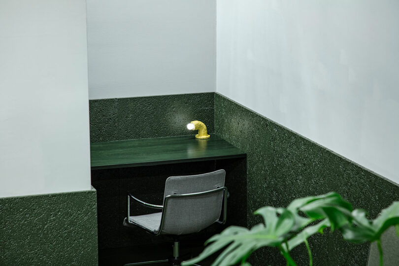 A small workspace with a green desk, a gray chair, and a yellow pipe-shaped lamp in the corner of a room with green and white walls. A plant is visible in the foreground.