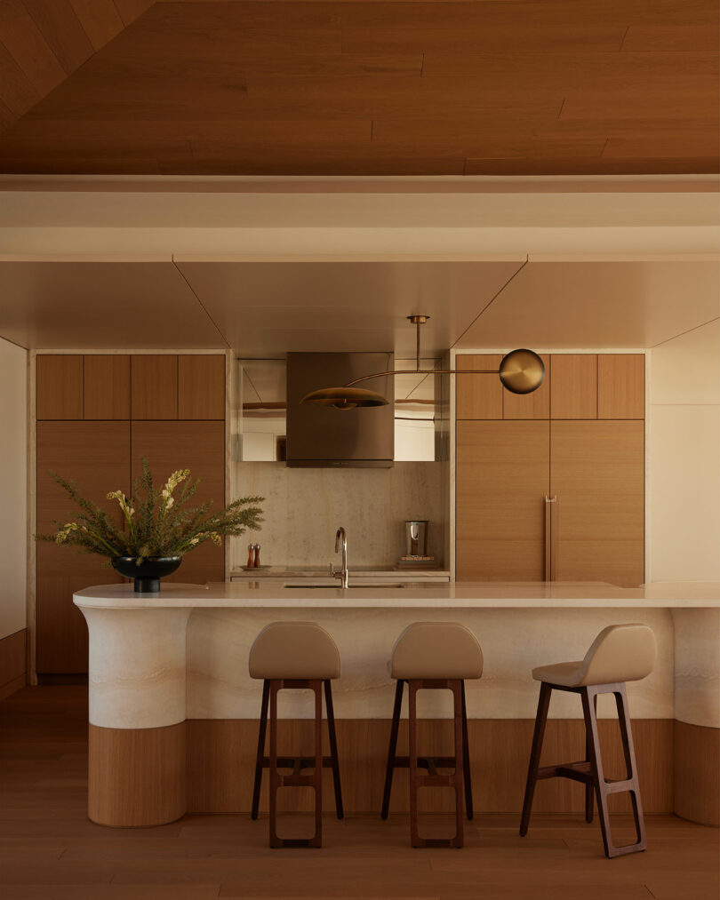 A modern kitchen with wood cabinets, a marble island with three beige stools, a built-in oven, and a vase of greenery on the counter.
