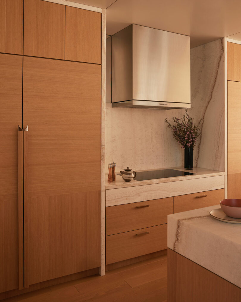 Modern kitchen with light wood cabinets, built-in refrigerator, marble countertops and backsplash, induction cooktop, stainless steel hood, and a vase with flowers.