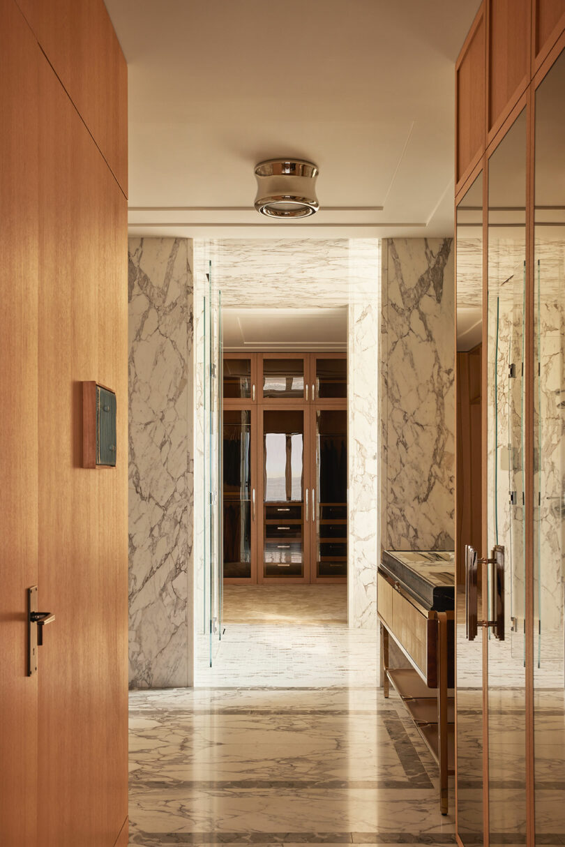 A hallway with marble floors and walls, wood-paneled doors, glass accents, and a mirrored console table, leading to a room with wooden shelving.