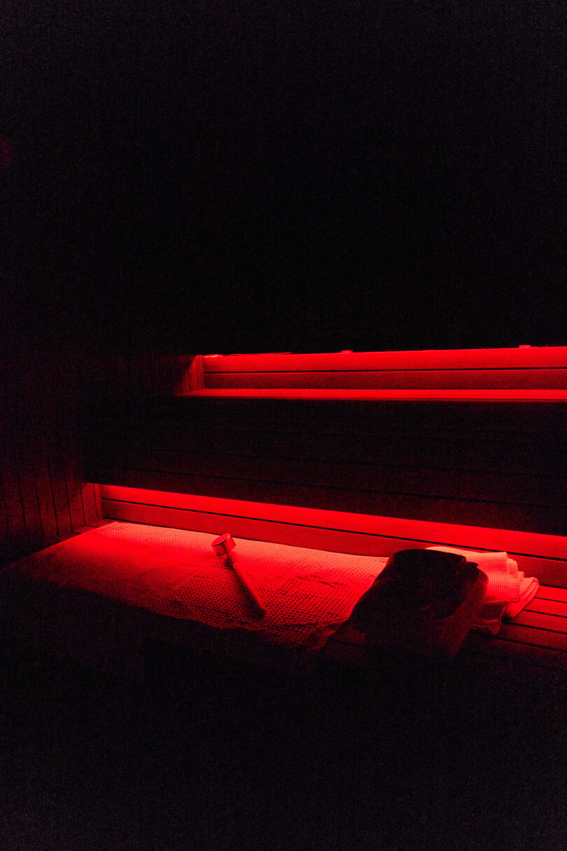 A dimly lit sauna interior features red lights, a wooden bench with a towel, and a ladle resting on top.