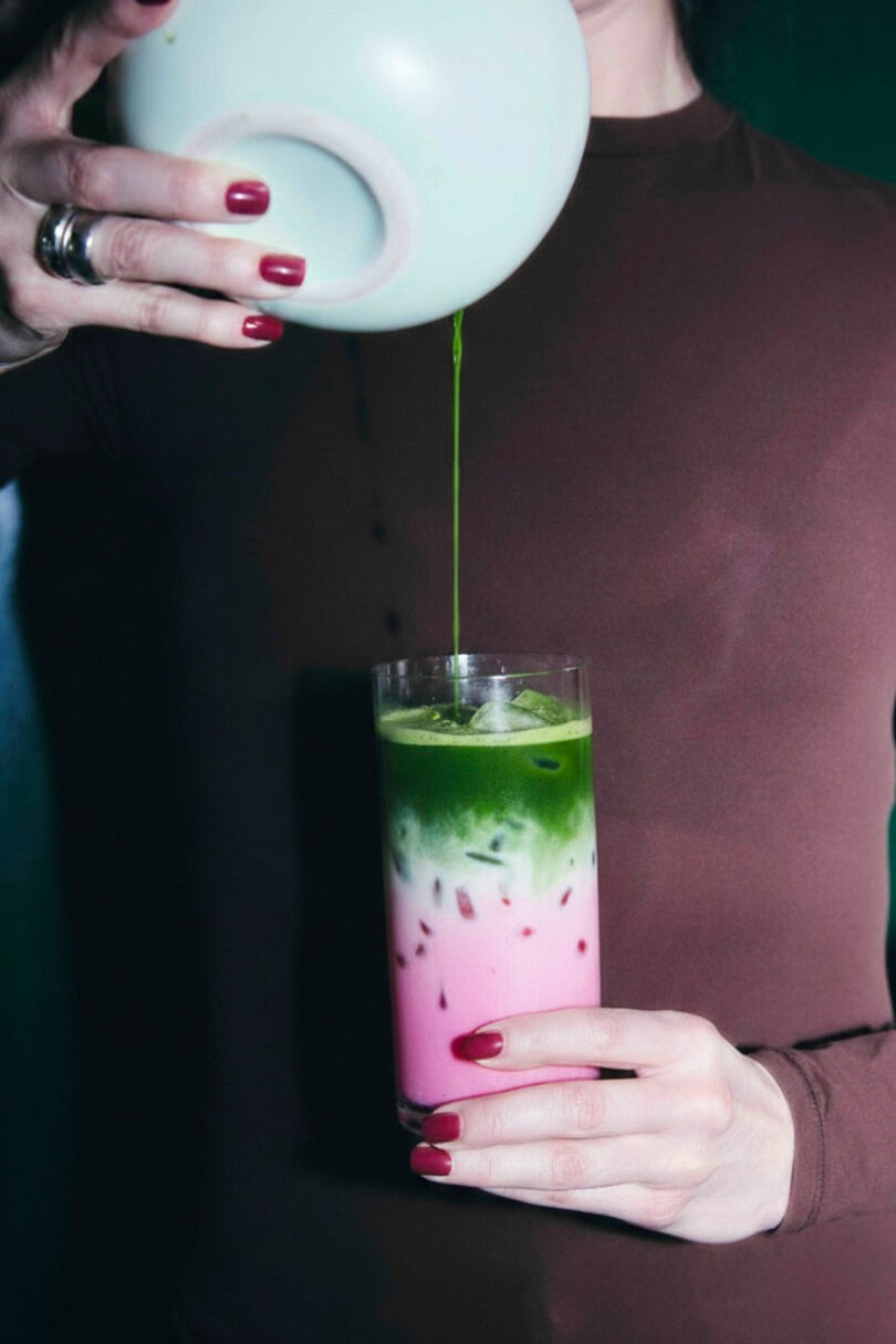 A person pours a green liquid from a bowl into a tall glass of iced pink and green layered drink, holding the glass with one hand.