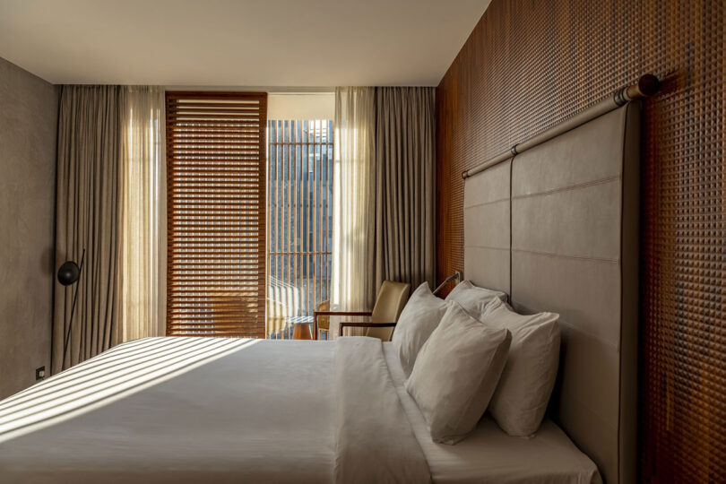 Hotel room with a large bed, four white pillows, a wooden headboard, a chair, and a small desk—sunlight streams through wooden blinds onto the bed, highlighting interior details inspired by Malik Architecture.