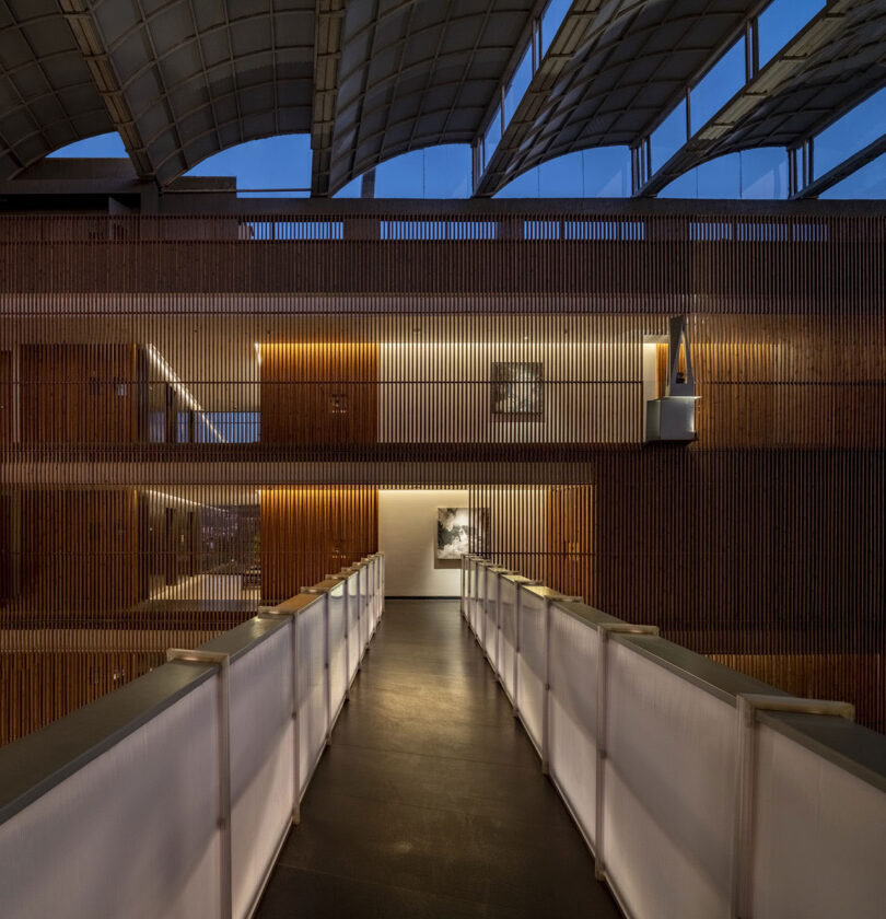 An illuminated indoor walkway with translucent railings leads to a modern space by Malik Architecture featuring wood paneling and a high, curved ceiling at dusk.