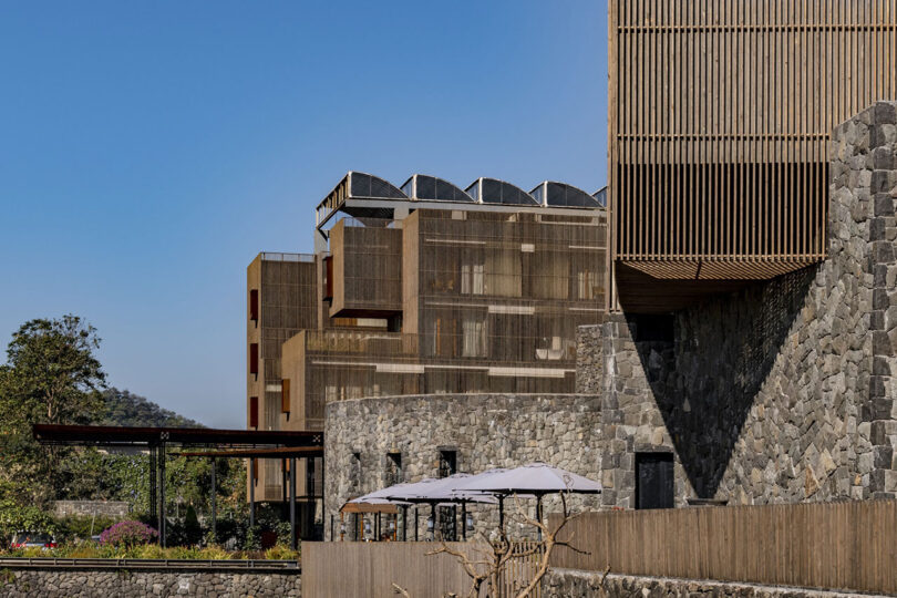 Modern multi-story building by Malik Architecture with wooden slats and stone walls, featuring outdoor umbrellas and mountains visible in the background under a clear blue sky.