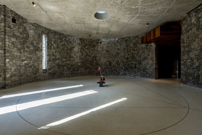 A circular stone-walled room designed by Malik Architecture, featuring a central skylight, narrow vertical window, and a red robotic arm poised in the center of the concrete floor.