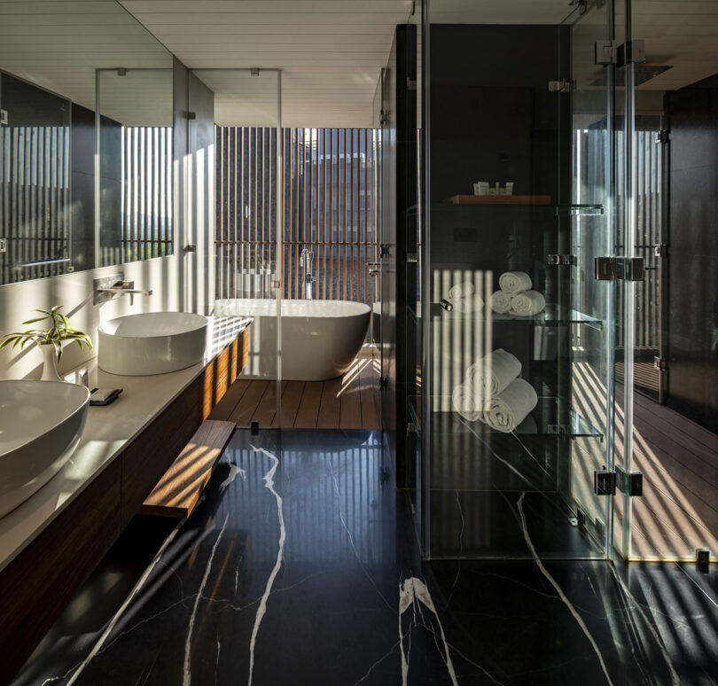 Modern bathroom by Malik Architecture with glass shower, vessel sinks, a freestanding bathtub, wooden accents, and sunlight streaming through vertical slats, creating striped light patterns on surfaces.