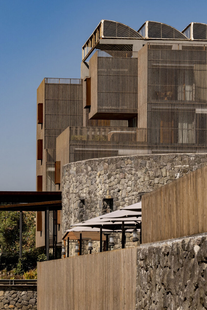 Modern building by Malik Architecture with vertical wooden slats and geometric sections sits above a stone wall with a terrace and white umbrellas, set against a clear blue sky.