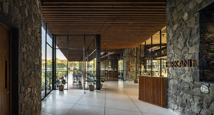 Modern restaurant entrance designed by Malik Architecture, featuring floor-to-ceiling glass walls, stone accents, wooden ceiling beams, potted plants, and a host stand labeled "ORKAN." Outdoor seating is visible through the windows.