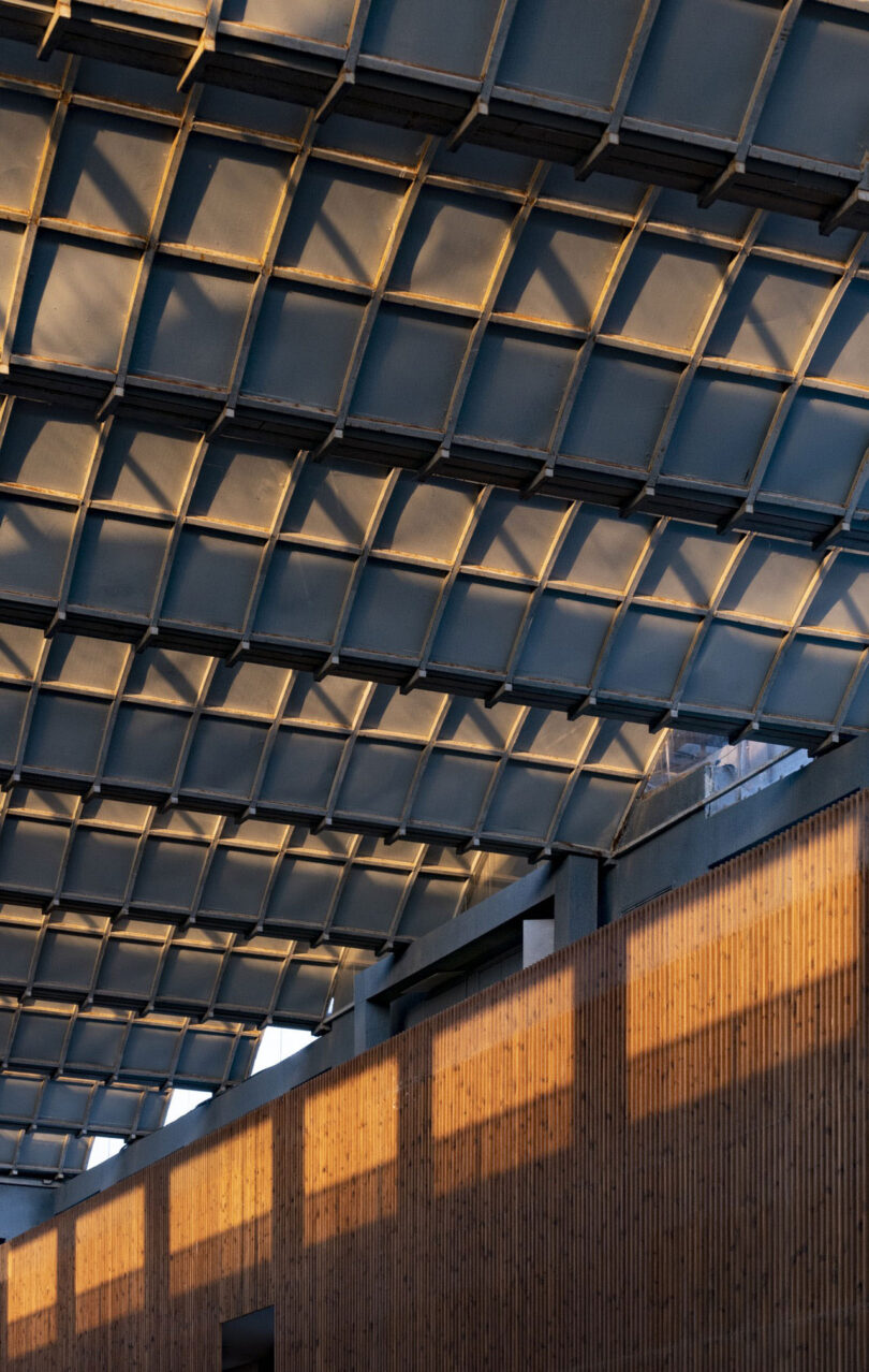 A geometric metal roof by Malik Architecture casts shadows on a wooden wall below, with warm sunlight highlighting the structure's repetitive grid pattern.