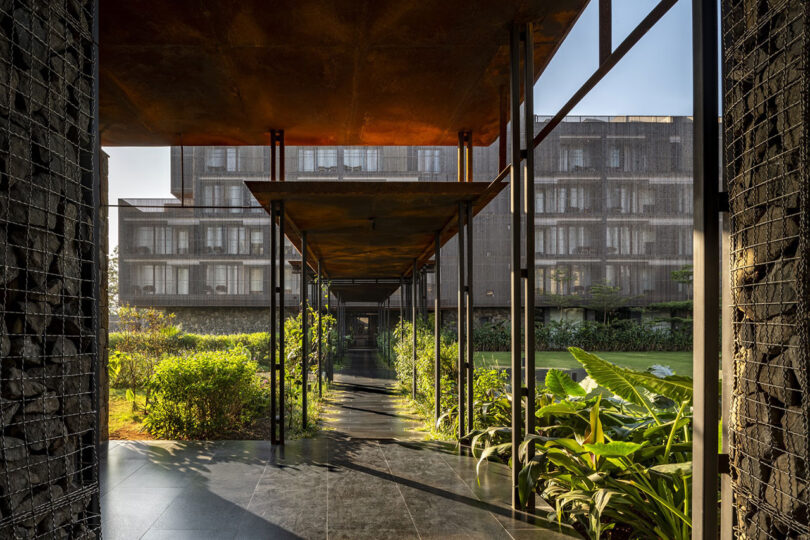 Covered walkway with metal roof panels and supports, surrounded by greenery, leading towards a modern, multi-story building in the background—showcasing the signature style of Malik Architecture.