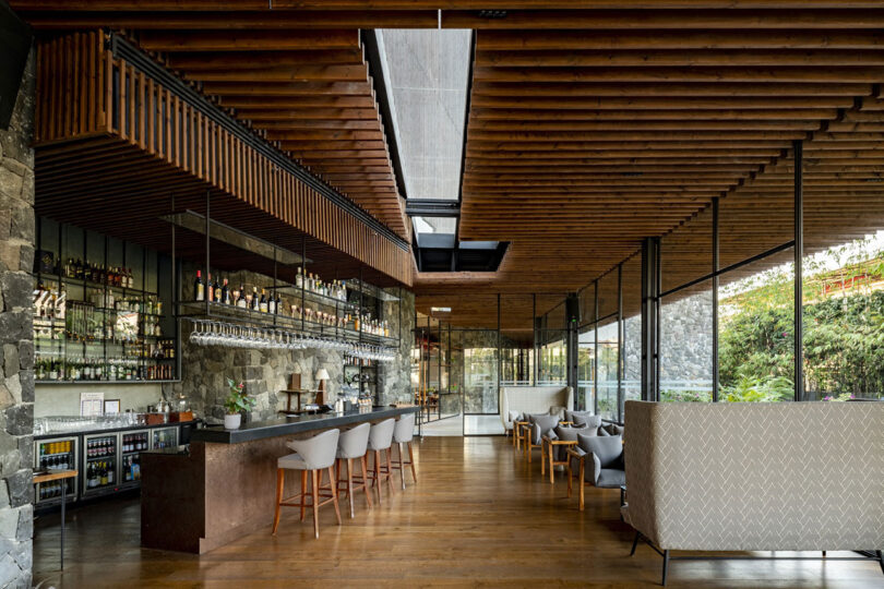 Modern bar and lounge area by Malik Architecture, featuring stone walls, wooden ceiling slats, a long bar with stools, glass shelves of bottles, and upholstered chairs on a wooden floor. Large windows provide natural light.