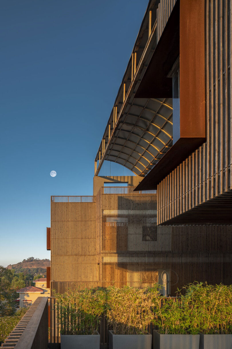 A modern building by Malik Architecture with a rust-colored metal facade and vertical slats is shown at sunset, potted plants in the foreground and the moon visible in the clear sky.