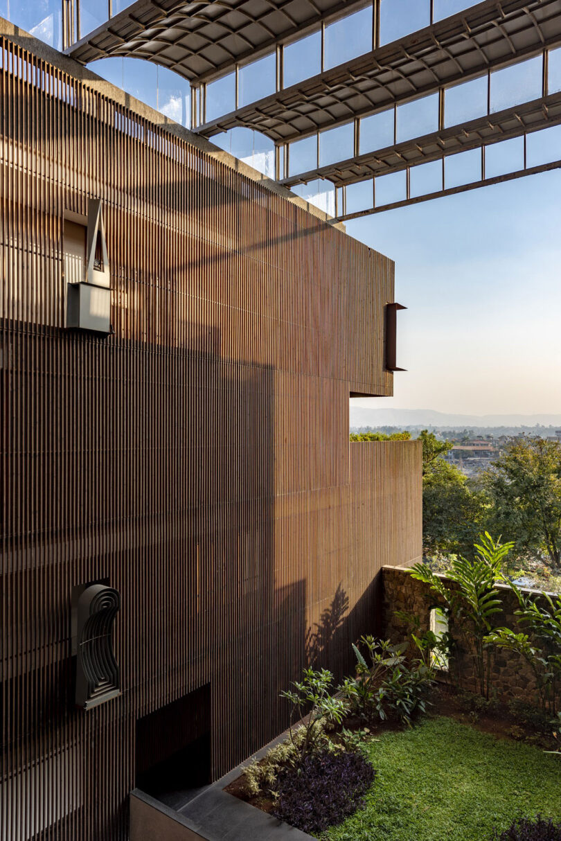 A modern building by Malik Architecture with a wooden slatted exterior stands beside a small garden, under a large overhanging roof structure, offering landscape views in the background.