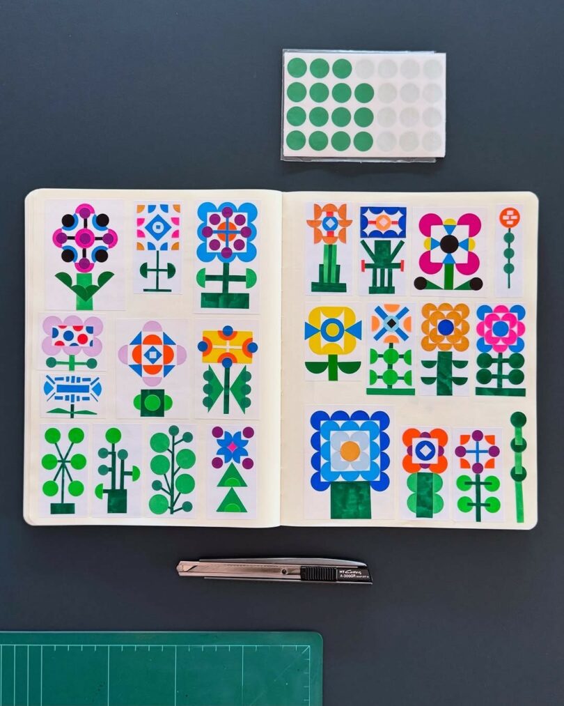 An open notebook displays colorful geometric flower designs made with circle stickers, reminiscent of a Design Milk Take 5 feature, with a sheet of green stickers, a utility knife, and a green cutting mat arranged nearby on a dark surface