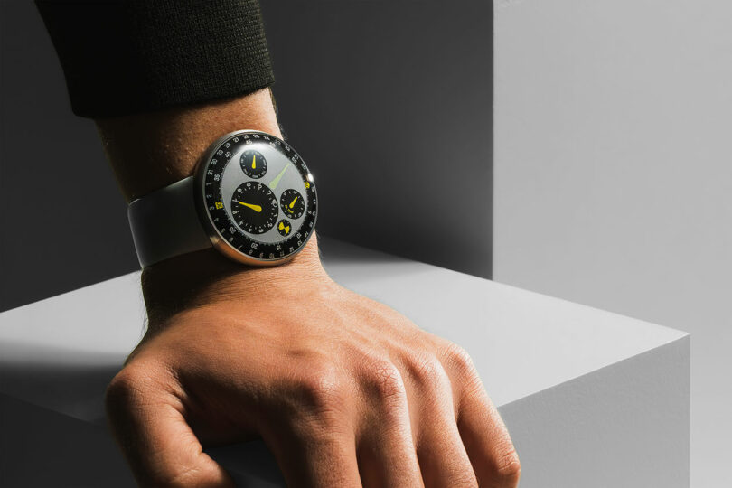 A person's wrist wearing a modern TYPE 3 analog watch with a black band and multiple subdials, resting on a gray geometric surface.