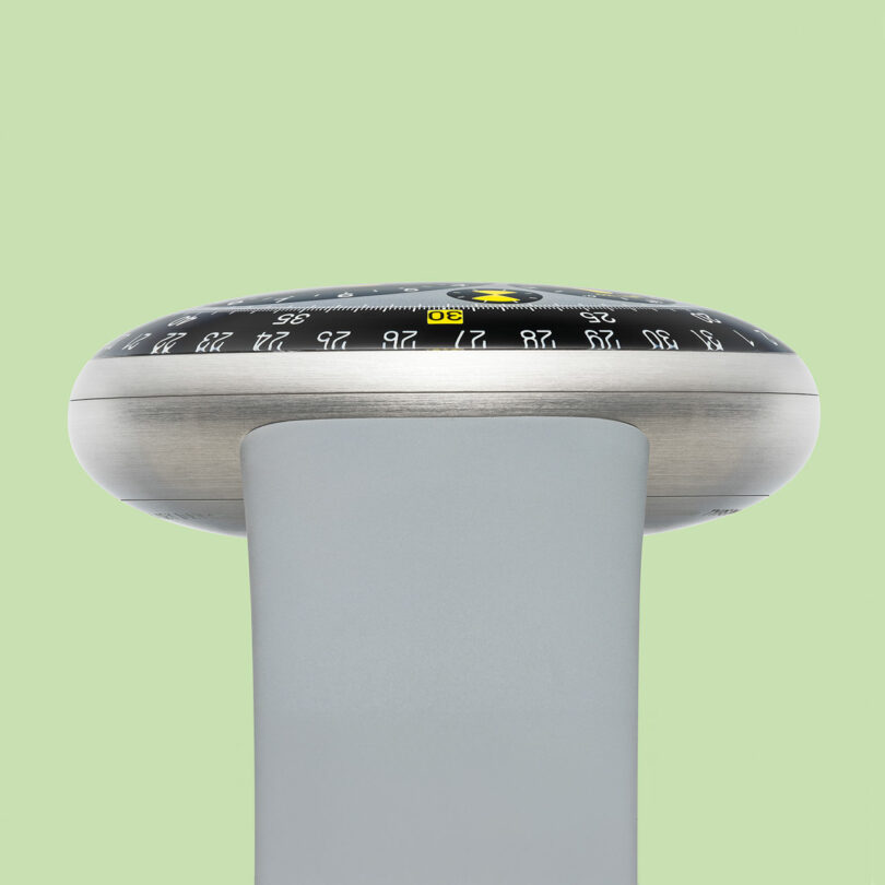 A modern TYPE 3, disc-shaped analog watch with black and silver detailing sits atop a smooth gray stand against a light green background.
