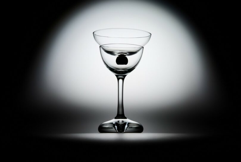 A clear, empty glass with a thin stem is centered against a white-to-black gradient background, lit dramatically from behind