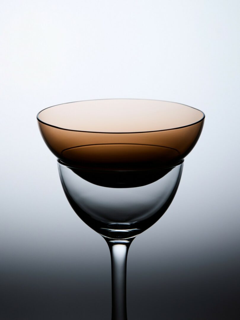 A clear glass cup with a wide, shallow, brown-tinted bowl stacked upside down on top of it, set against a light gradient background
