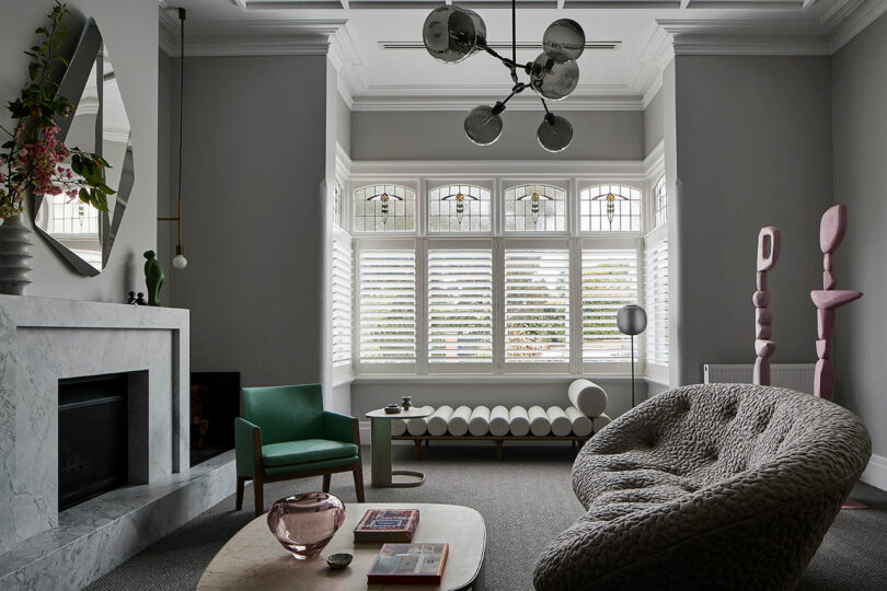 Modern living room with large shuttered windows, gray walls, contemporary furniture, a marble fireplace, abstract sculptures, and geometric light fixture.
