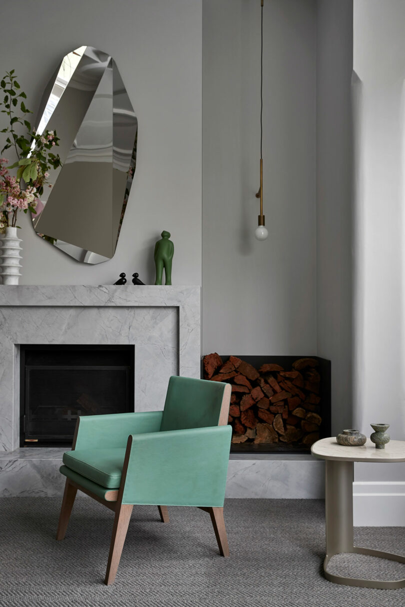 Modern living room with a mint green chair, marble fireplace, geometric mirror, stacked firewood, hanging bulb light, and minimalist decor.