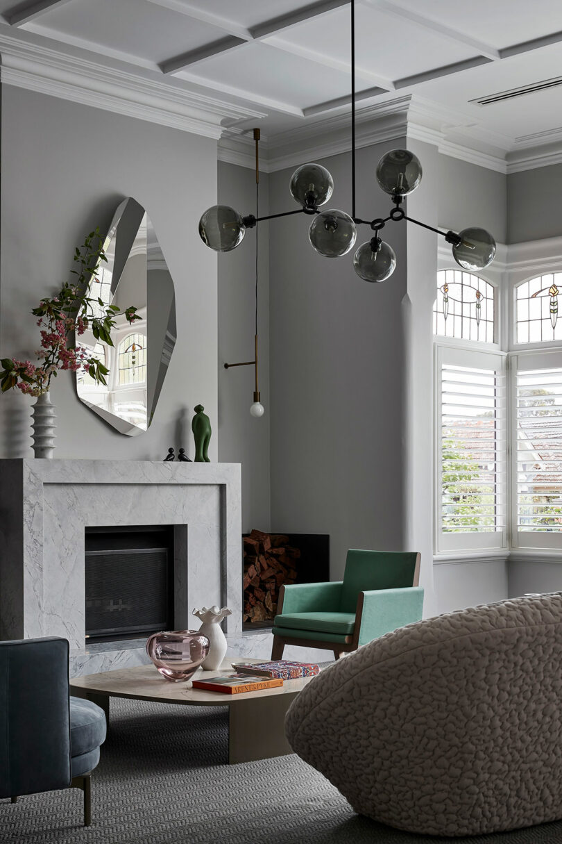 Modern living room with a marble fireplace, geometric mirror, contemporary chandelier, green armchair, textured sofa, coffee table, and shuttered windows.