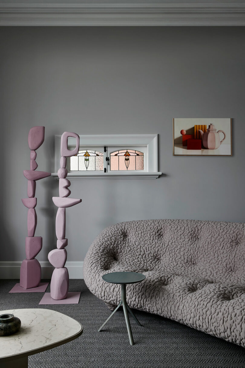 A minimalist living room with a textured grey sofa, small round side table, abstract pink sculptures, and a shelf with a framed artwork and two stained glass windows.