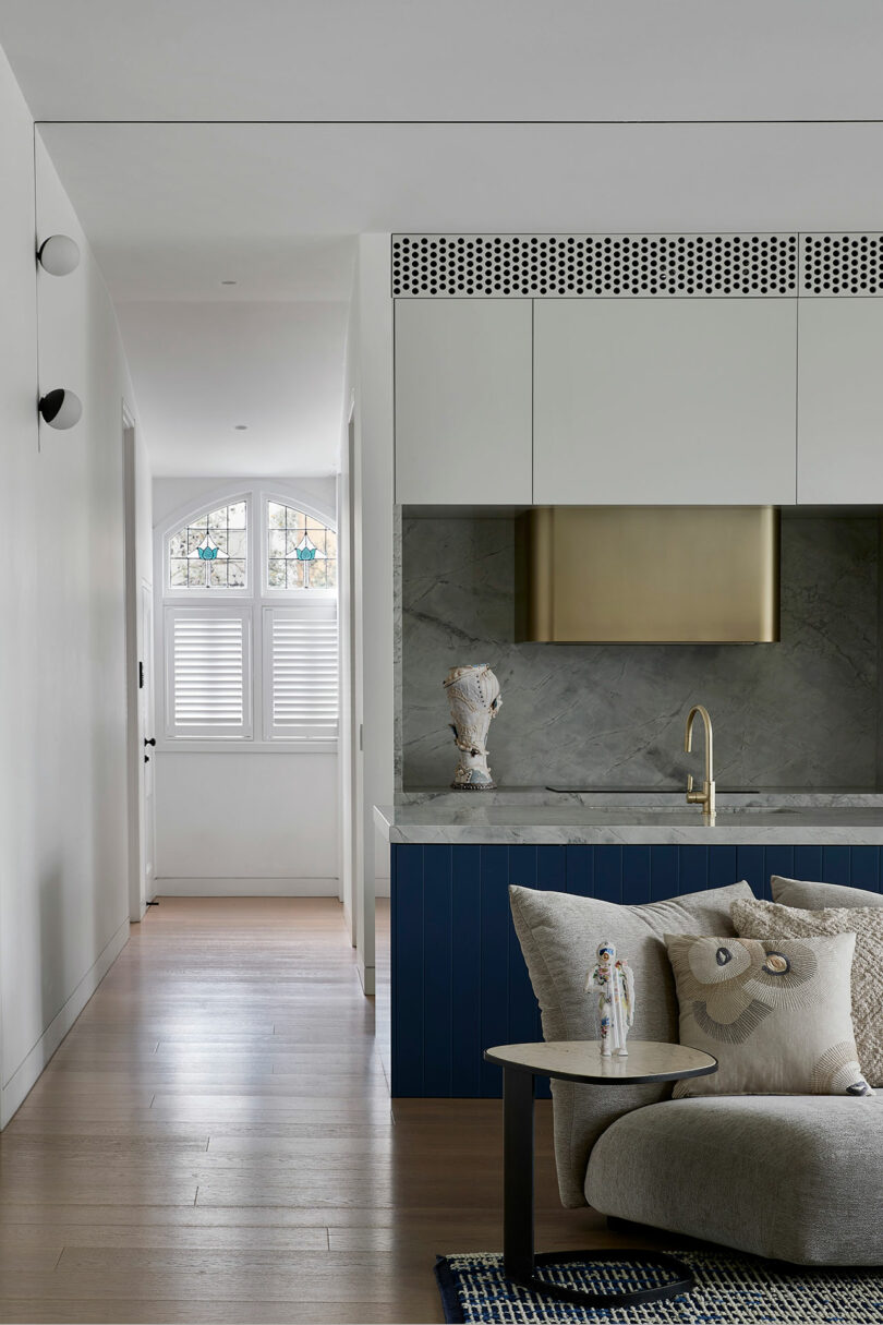 Modern living space with a blue kitchen island, brass range hood, marble backsplash, and decorative pillows on a curved sofa; hallway with wooden floors and a window with white shutters.