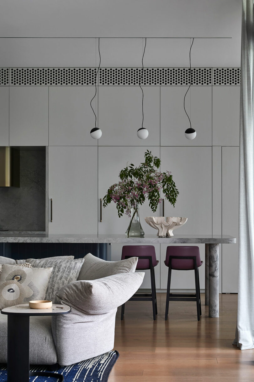 Modern living and dining area with white cabinetry, marble table, two purple chairs, pendant lights, and a vase with flowers on the table. Neutral tones and minimalist decor.