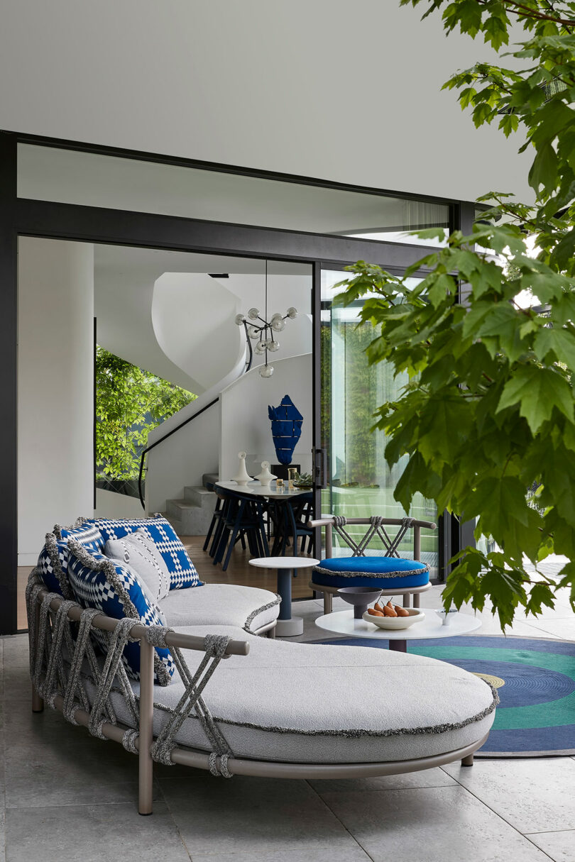 Modern patio with a curved lounge chair and blue-accented cushions, set beside glass doors leading to a dining area and a spiral staircase, with green leaves in the foreground.