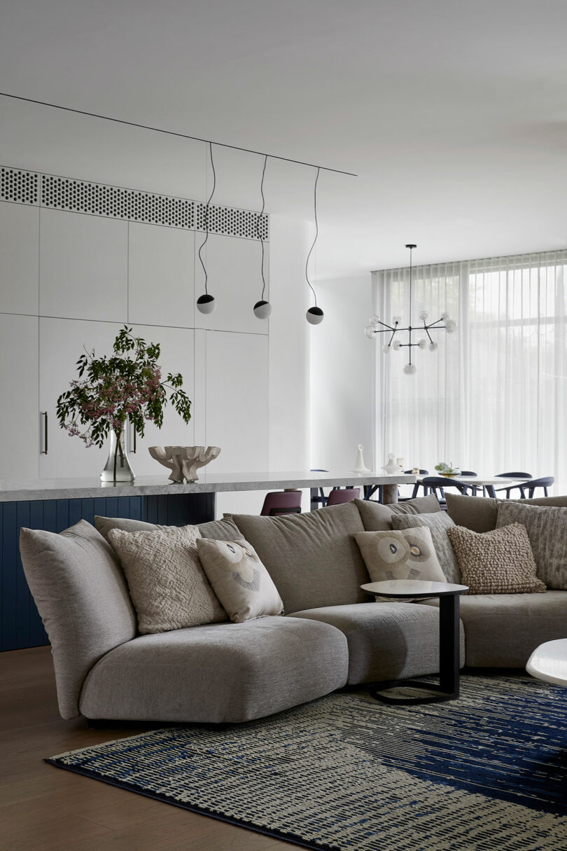 Modern living room and dining area with a gray sectional sofa, textured cushions, a blue-patterned rug, minimalist pendant lights, and large windows with sheer curtains.