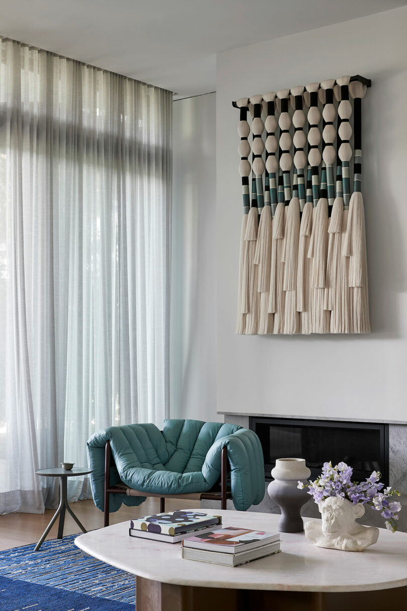 A modern living room with sheer curtains, a teal lounge chair, a marble coffee table with books and decor, and a large woven wall hanging above a fireplace.