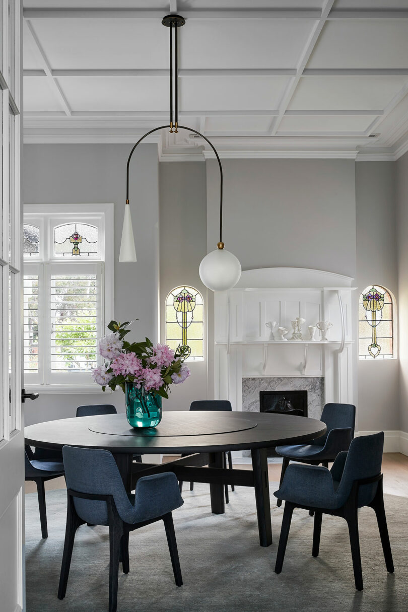 A modern dining room with a round black table, six blue chairs, a vase of pink flowers, a white fireplace, stained glass windows, and a contemporary pendant light.