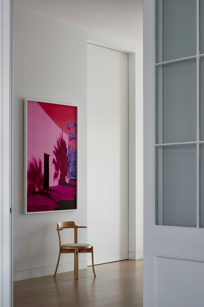 A wooden chair sits below a framed artwork with a pink architectural scene and palm shadows in a minimalist room with white walls and light wood flooring.