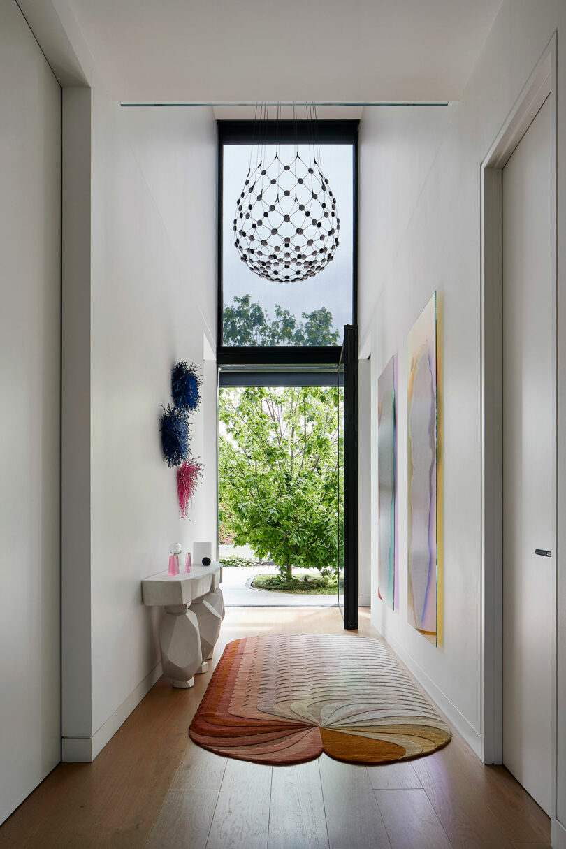 Modern hallway with large window, geometric chandelier, abstract wall art, a textured rug, sculptural console table, and a view of greenery outside.